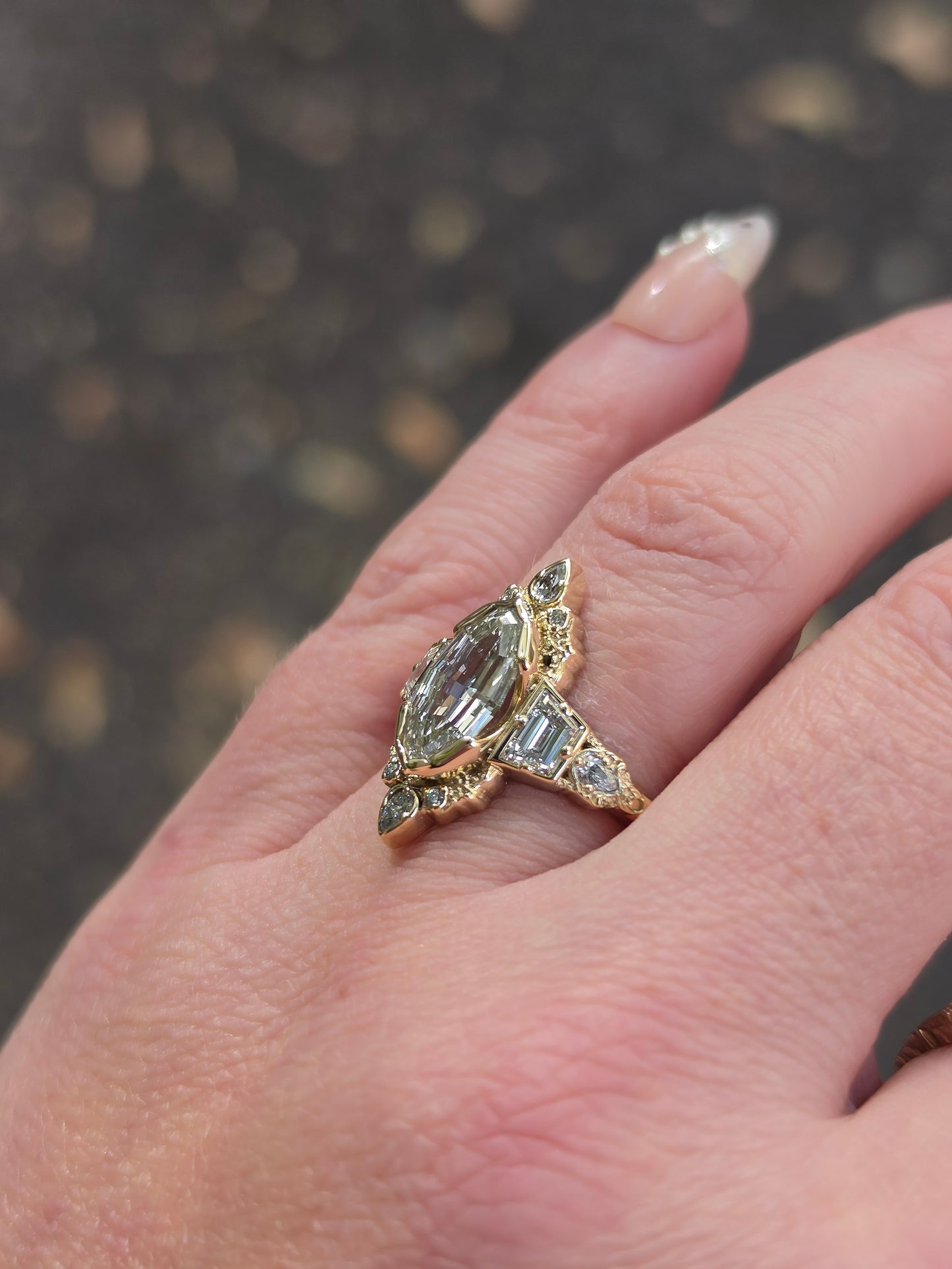 A close-up of a hand wearing a vintage-style gold ring with a marquise-cut gray-green center stone and baguette side stones, set against a blurred dark outdoor background.