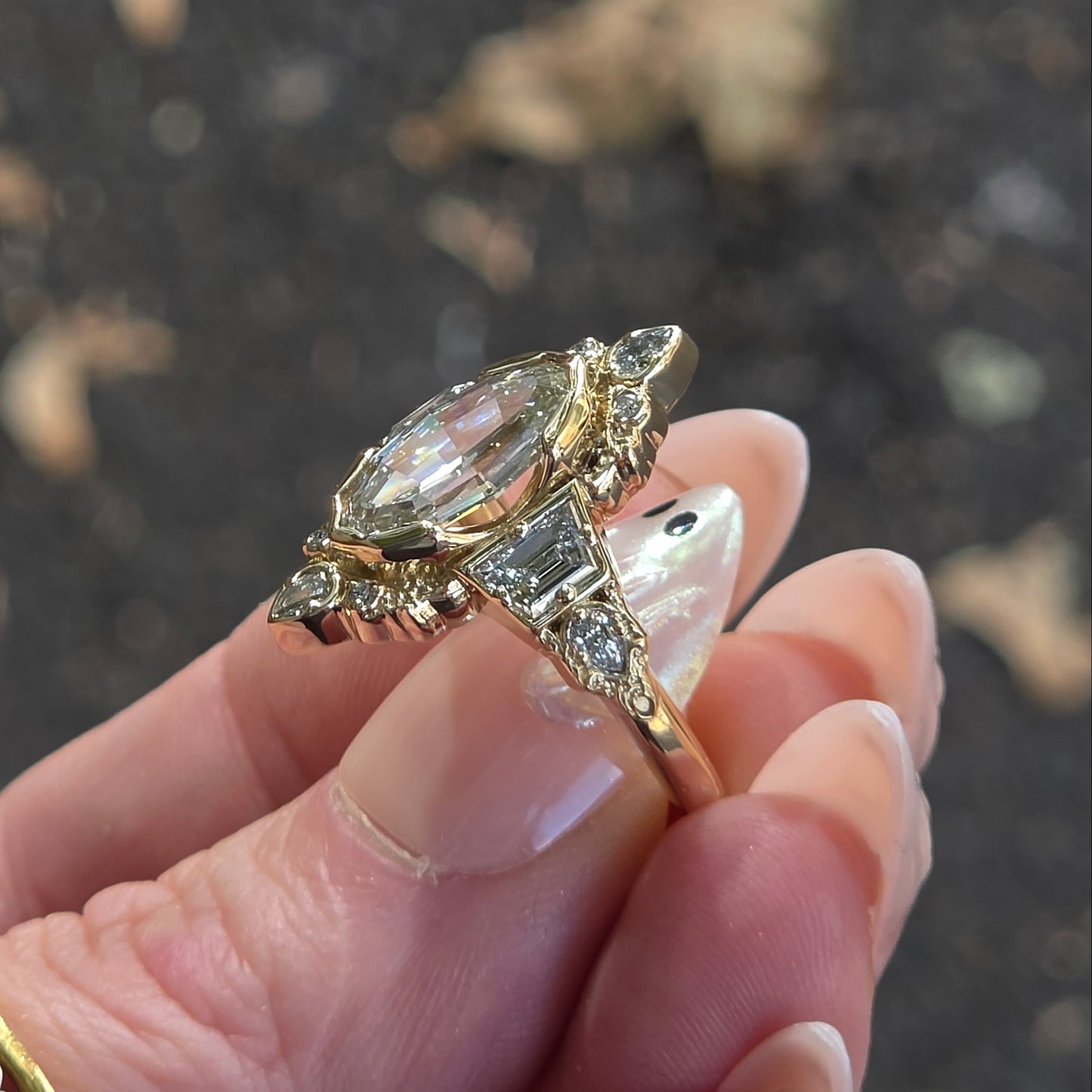 A gold ring with a large marquise-cut clear gemstone and smaller accent stones, held between fingers against a blurred dark outdoor background.