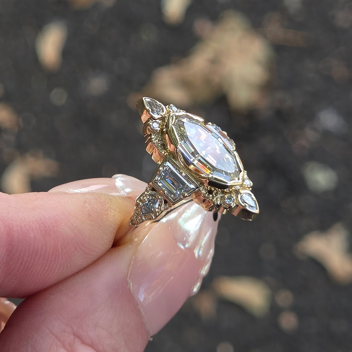 A gold ring with a large marquise-cut clear gemstone and smaller accent stones, held between fingers against a blurred dark outdoor background.