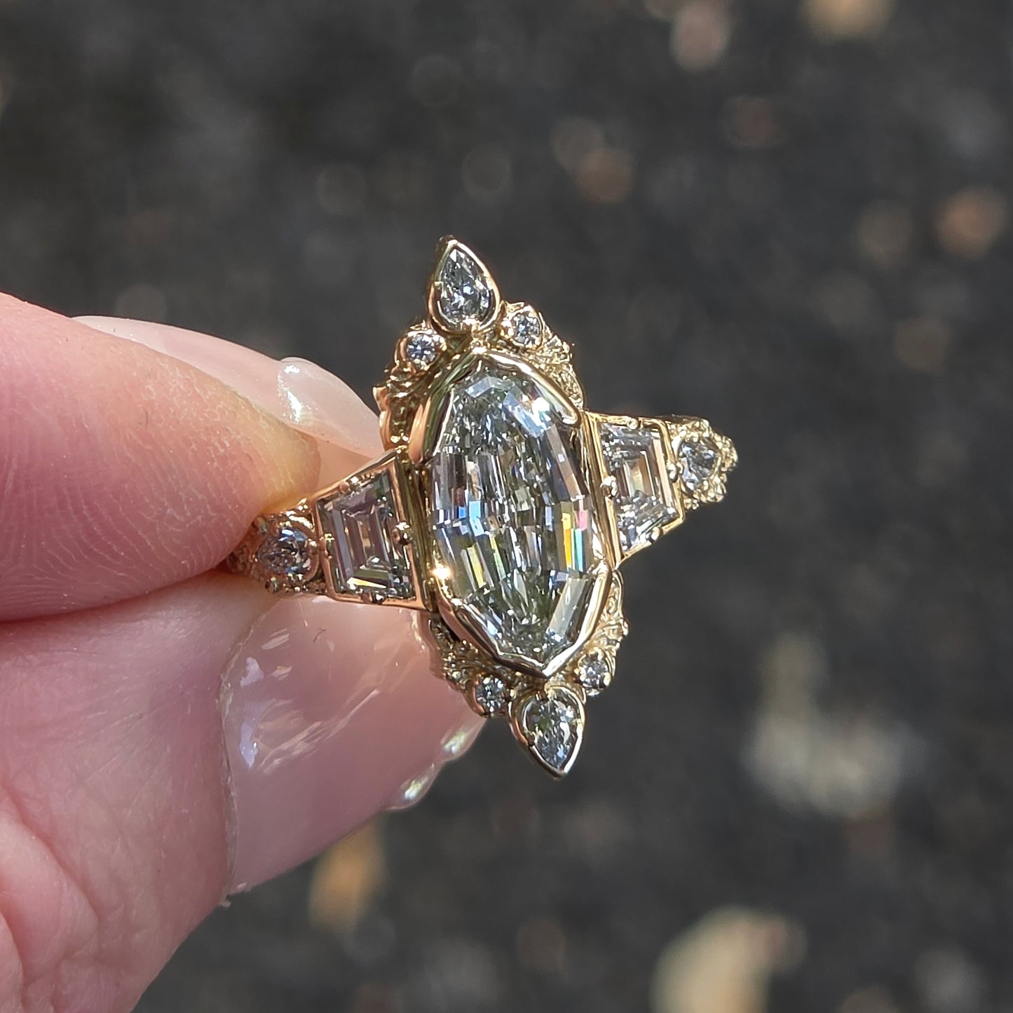 A gold ring with a marquise-cut clear center stone and smaller accent stones, held between fingers against a blurred dark outdoor background.