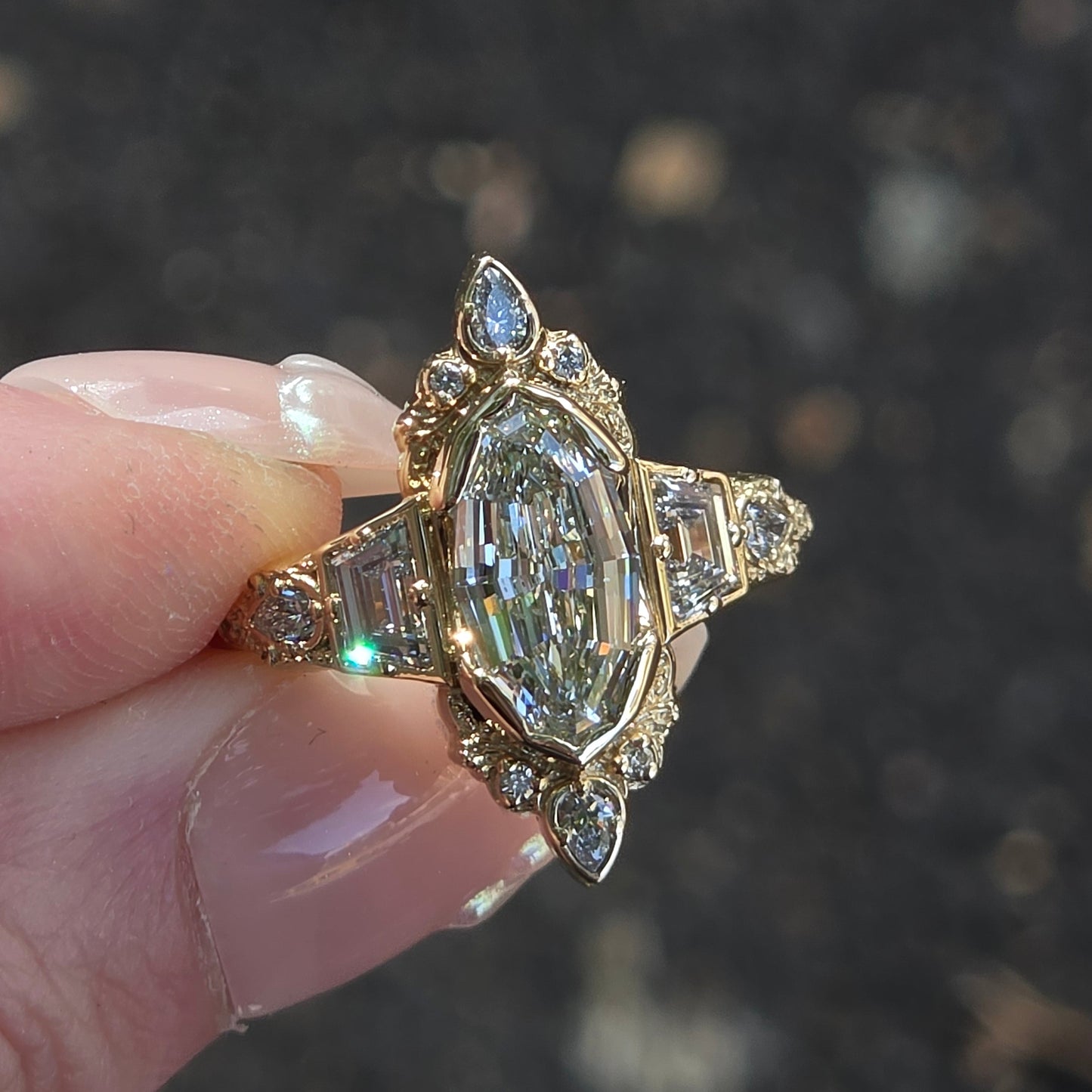A close-up of a hand holding a vintage-style gold ring with a large marquise-cut clear center stone and smaller accent stones, set against a blurred dark outdoor background.