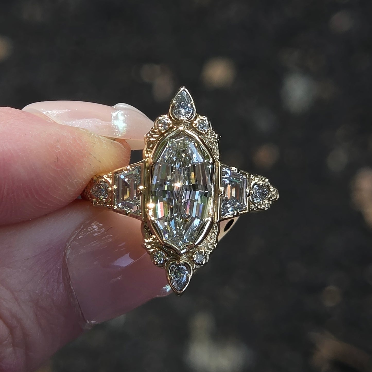 A vintage-style diamond ring with a marquise-cut center stone and smaller diamonds on the band, held between fingers against a dark, blurred outdoor background.