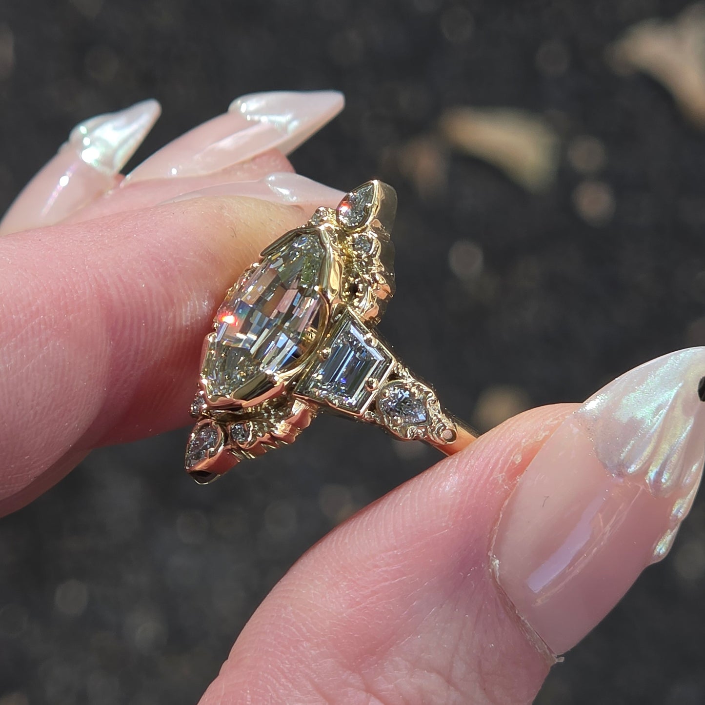 A close-up of a hand with long manicured nails holding a vintage-style ring. The ring features a gold-toned setting with a marquise-cut clear gemstone and baguette-cut clear gemstones, set against a dark, blurred outdoor background.