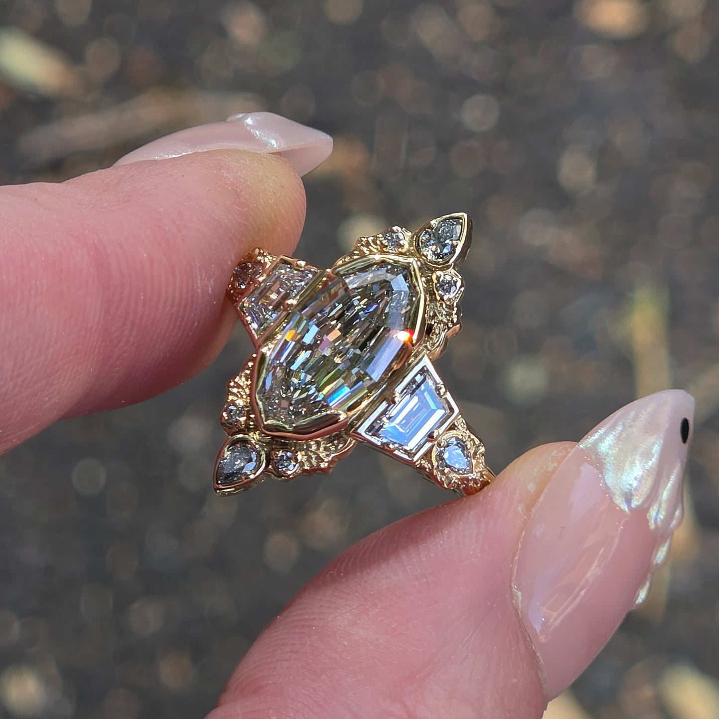 A gold ring with a large marquise-cut clear center stone and smaller clear accent stones, held between fingers against a blurred outdoor background.
