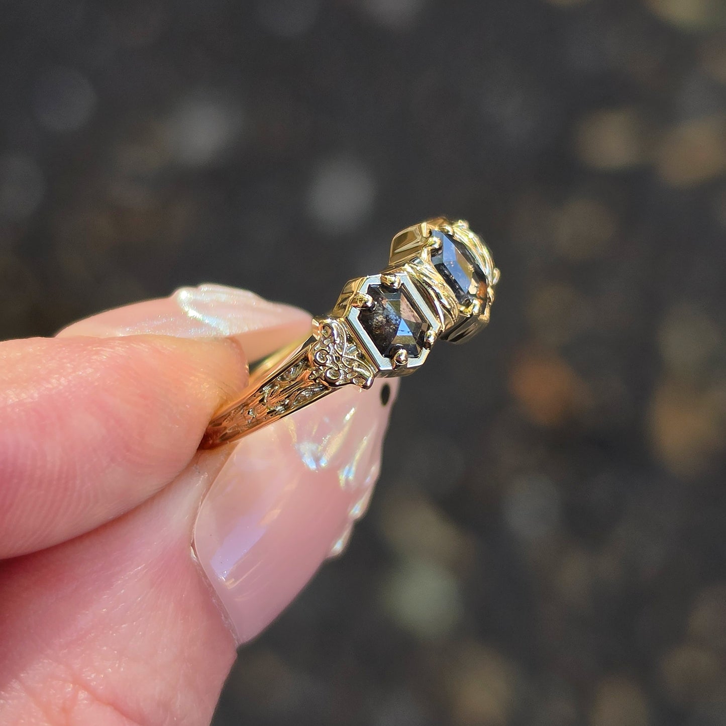 A close-up of a hand holding a yellow gold ring with three duchess-cut dark gray stones and small accent diamonds, set against a blurred dark outdoor background.