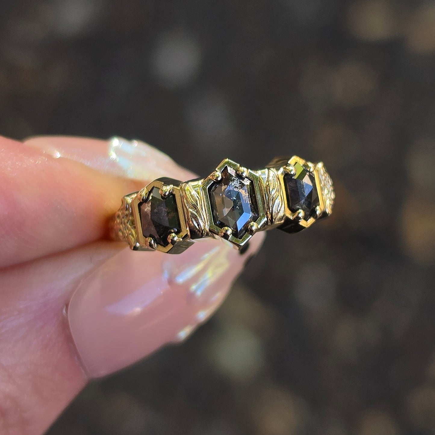 A gold ring with three duchess-cut dark gray stones, held between fingers against a blurred dark background.