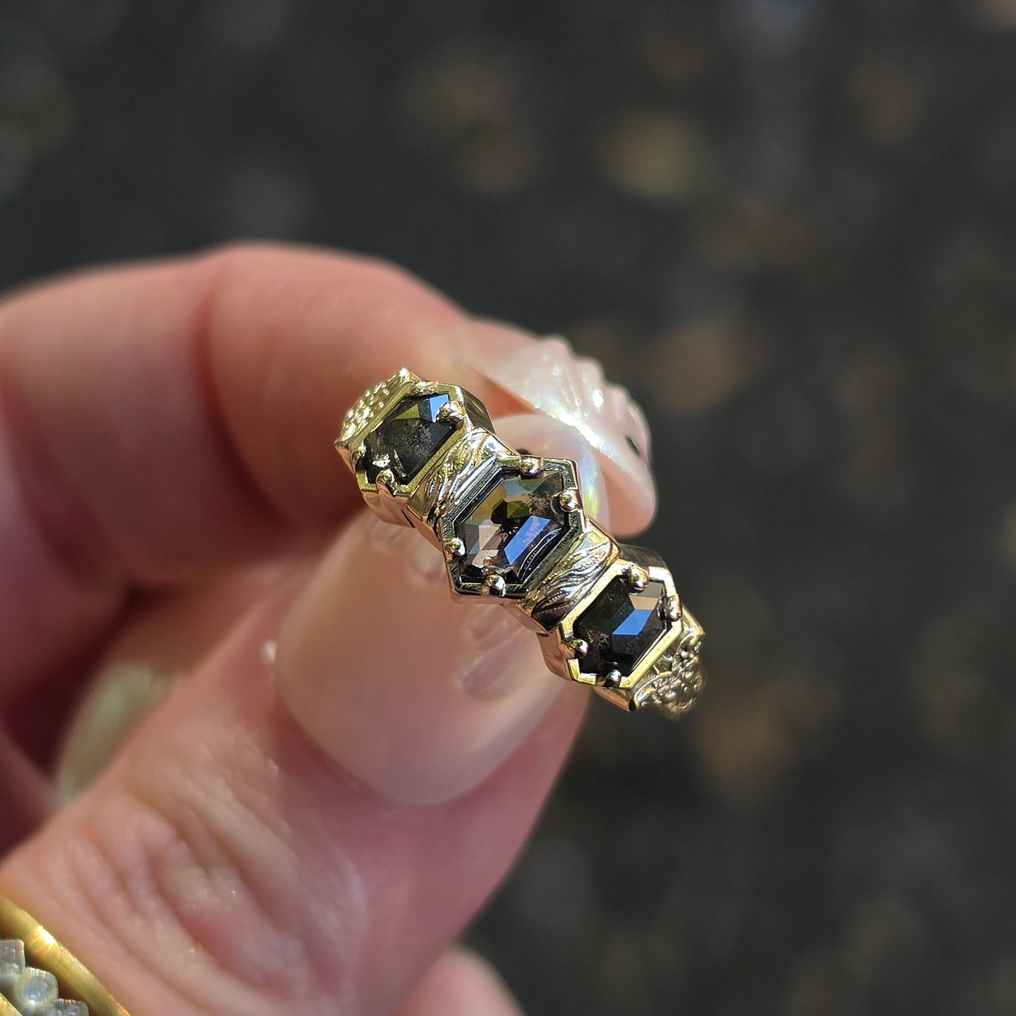A close-up of a hand holding a yellow gold ring with three hexagon-cut dark  gemstones, set against a dark, blurred background.