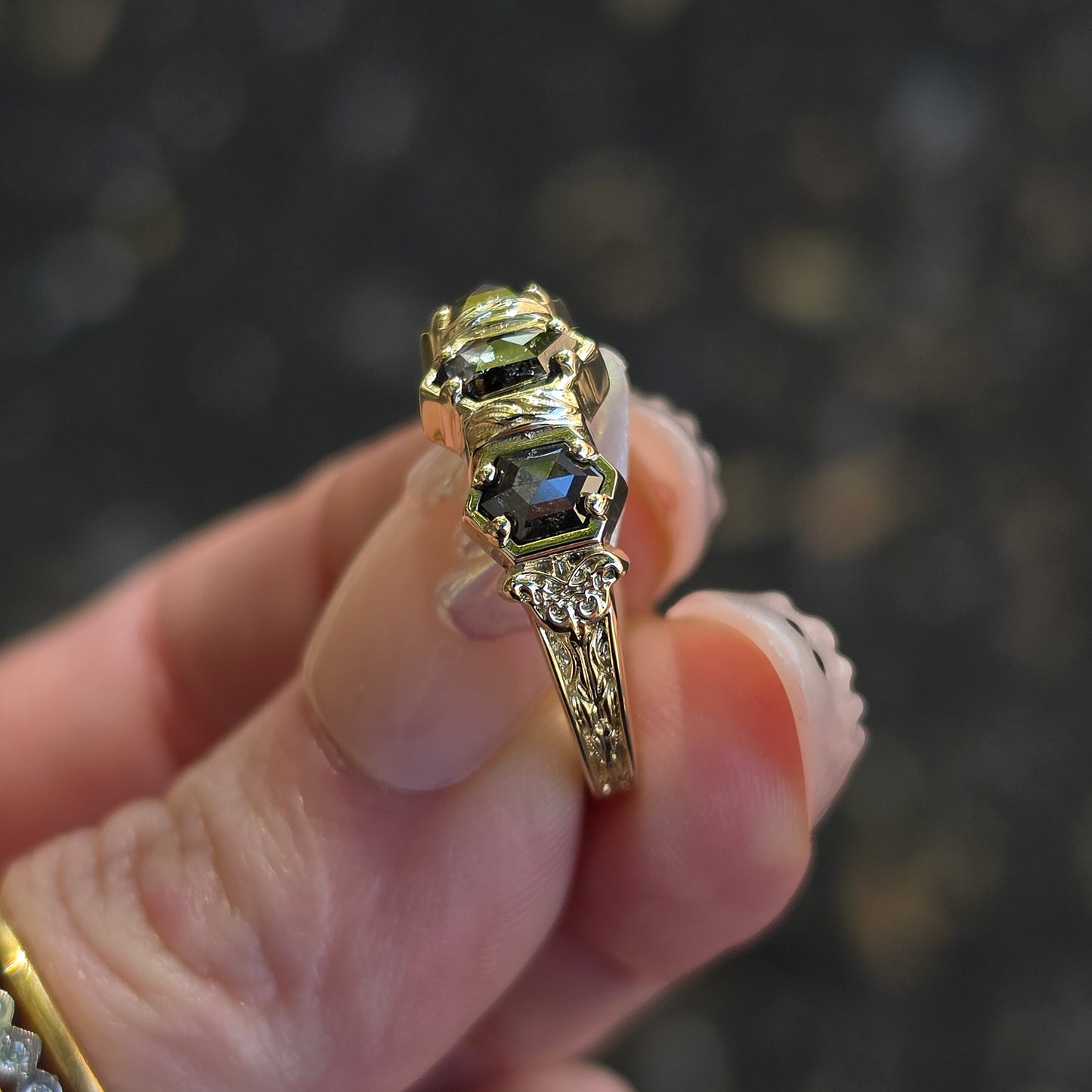 A gold ring with hexagon-cut salt pepper diamond gemstones held between fingers against a dark, blurred background.