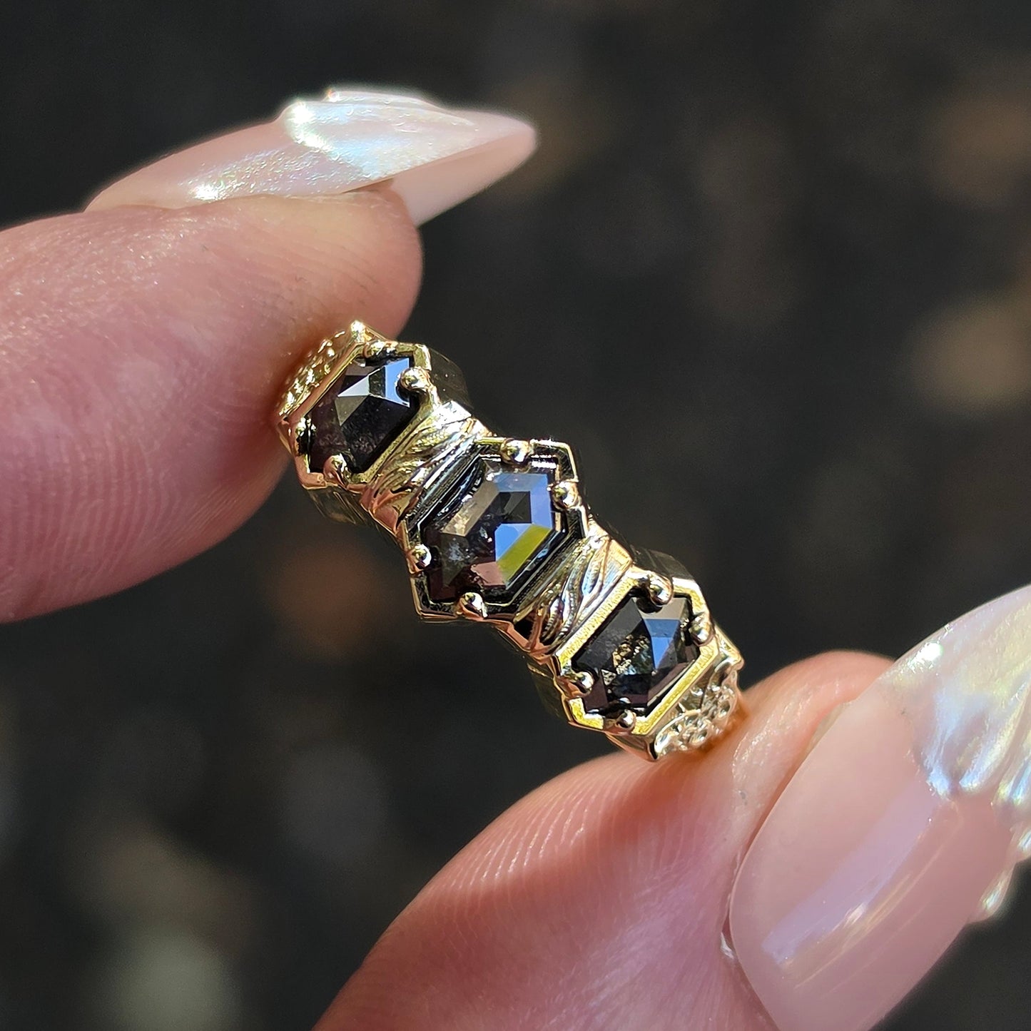 A gold ring with four dark iridescent gemstones held between fingers with long, manicured nails against a dark, blurred background.